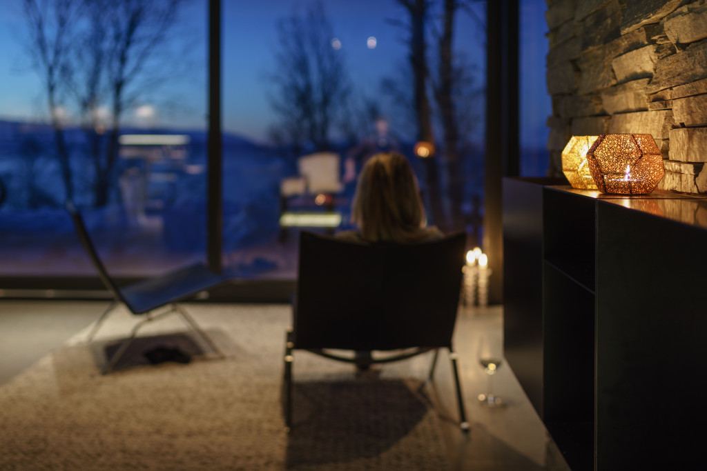 En kvinne sitter i en stol foran store vinduer i hytta i Russelv. Ute er det blå time over fjorden, inne lyser en lampe varmt.