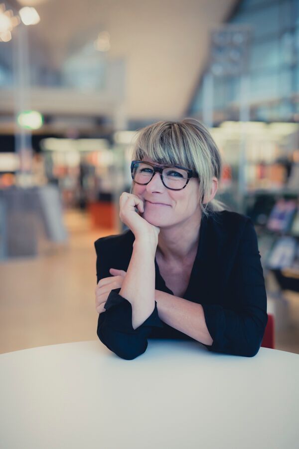 Torunn Tøllefsen smiler med haken i hånden, ved et hvitt bord i et lyst bibliotek.