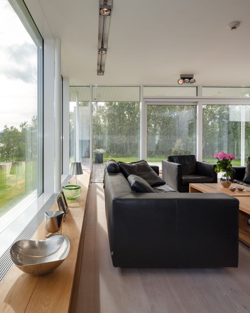 Stue i Villa Sirilund med sorte skinnsofaer foran vindusfelt fra gulv til tak og trær utenfor.